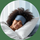 Close-up portrait of a young woman with dark, curly hair, smiling peacefully with her eyes closed, wearing a sleep mask.